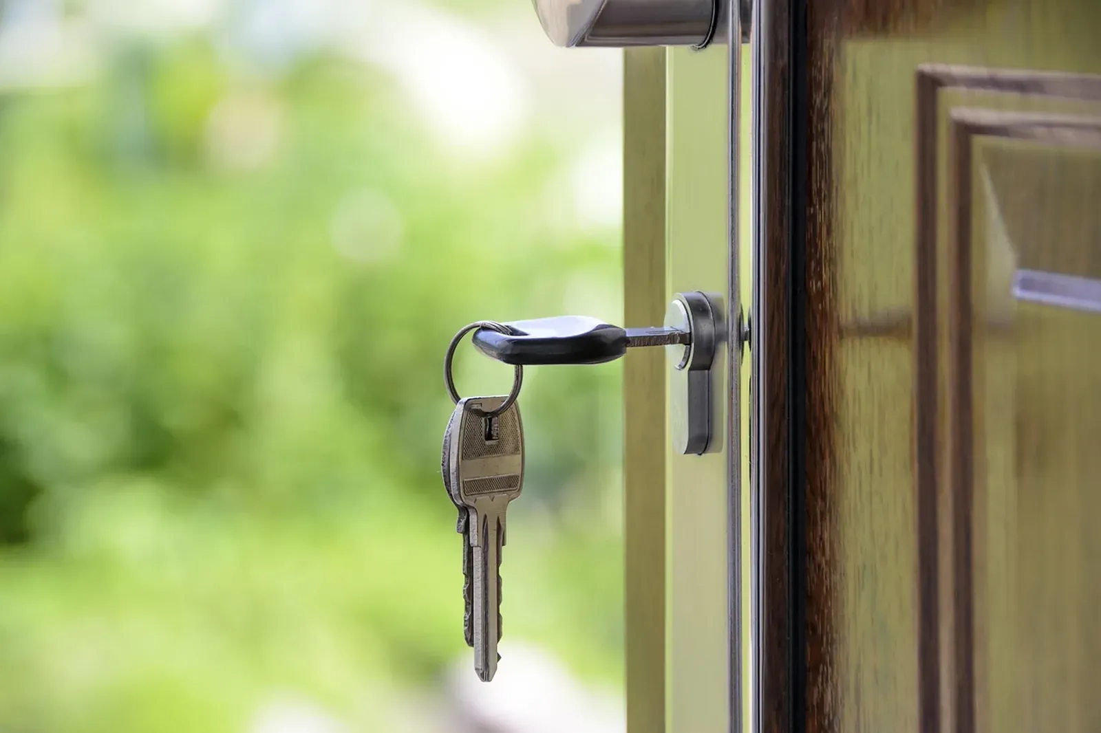 Set of house keys hanging from a cylinder lock on a wooden front door with green blur behind