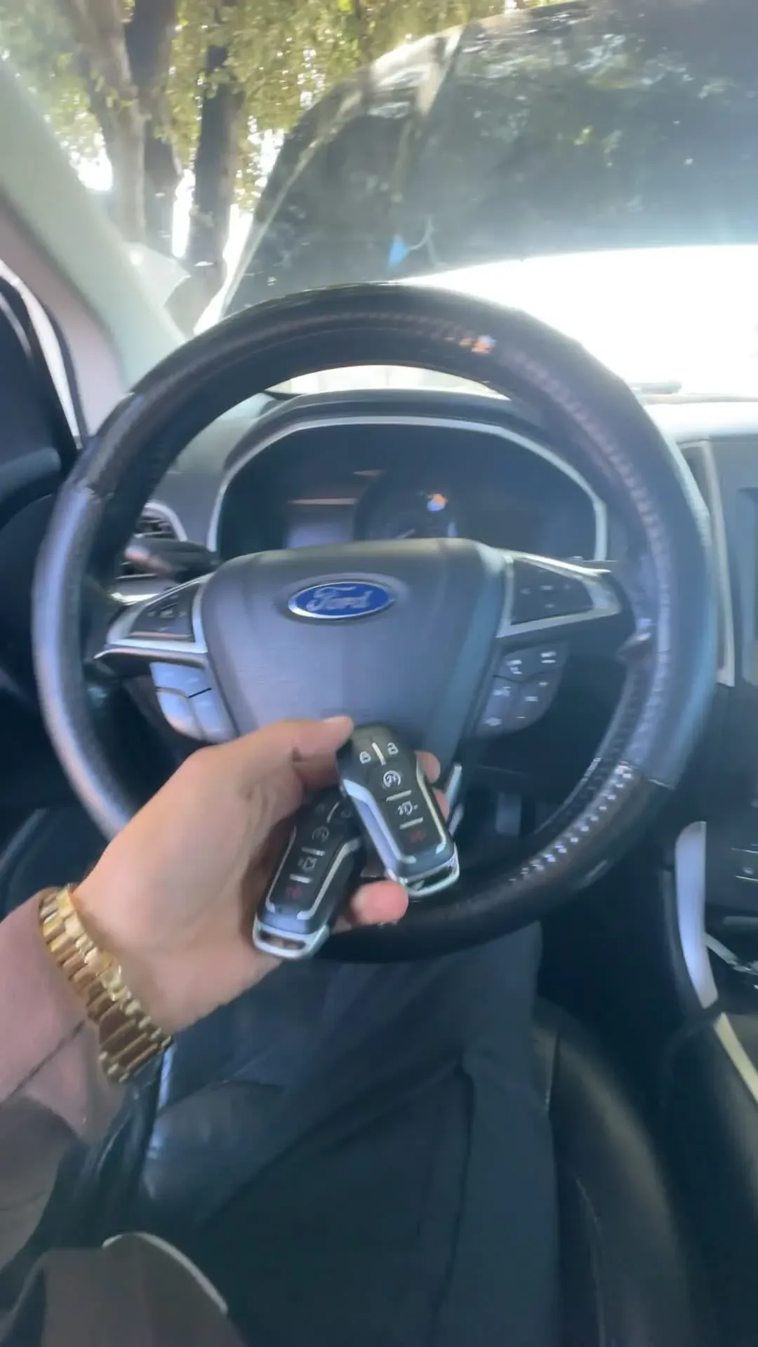 A hand holding several key fobs in front of a car steering wheel